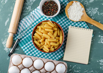 Fototapeta na wymiar Pasta in a bowl with eggs, starch, peppercorns, whisk, rolling pin, copybook top view on grunge and kitchen towel background