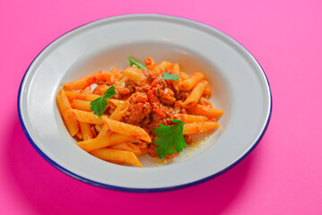 Plate of penne pasta with tomato sauce served in a white plate over bright pink background. Copy space banner.