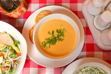 Pumpkin soup in a white bowl. Wooden table, red plaid tablecloth, blurred background. Dinner in restaurant.