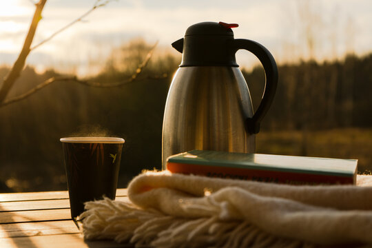 Thermos And A Hot Cup Of Tea Or Coffee With Book And Blanket While Social Distancing During Quarantine In Nature, Farm House Surrounded By Forest