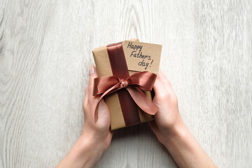 Womans hands holding gift box wrapped festive ribbon and label with text Happy Father's Day on wooden table, top view. Present for daddy, Fathers Day celebration concept.