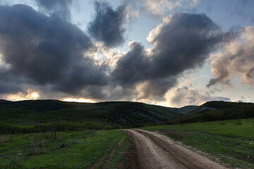 Landscape with a country road and clouds. Threatening rain clouds over the road. The sun breaks through the clouds. Rural spring landscape. A winding path leading into the forest.
