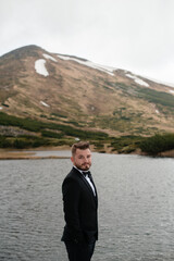 Wedding concept photo shoot. Beautiful young groom standing on a big stone in a lake and mountains with snow