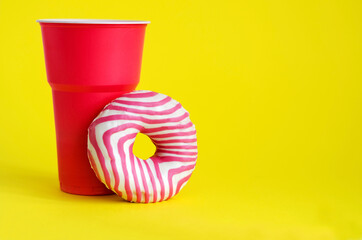 A donut stands next to a plastic pink glass on a yellow background. The concept of junk food, junk plastic dishes. Horizontal background, soft focus.