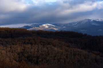 Panorama of snowy mountains on the background of an autumn forest. Snow peaks illuminated by the sun. Storm clouds hang over the mountain. Cold autumn landscape. Crimean mountains in January.