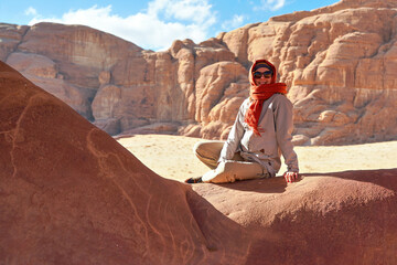 Fototapeta na wymiar Young woman wearing warm jacket and scarf around head smiling, sitting at naturally formed rock window in wadi rum desert on sunny day