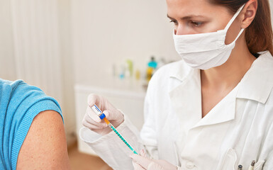 Female doctor in white coat and cotton face mask injecting vaccine into syringe, about to inject it...