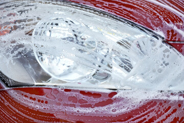 Front head light of red car washed in self serve carwash, white soap strokes on glass
