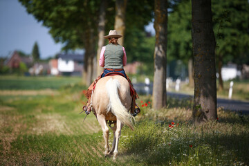 Cowboy rides along the street with a Palomino western horse, photographed from behind..