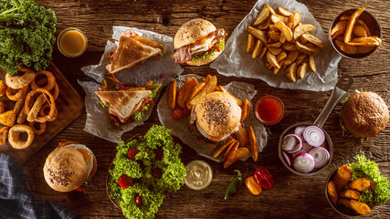 Top view of a table full of fast food, including hamburgers, potato chips, club sandwich, chicken nuggets and onion rings on a rustic table