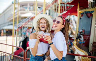 Obraz na płótnie Canvas Two young beautiful girls have fun in an amusement park. Drink lemonade and laugh. ladies enjoying weekend together