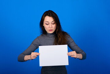 Fototapeta premium Young woman in grey shirt holding a sheet of paper and looking at the camera, smiling. Blue background isolated