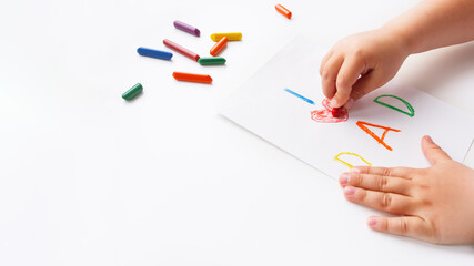 Toddler draws greeting card for Father's day. Kid uses wax crayons to write I Love Dad and to paint red heart symbol. Child's hands and pencils on white background with copy space.