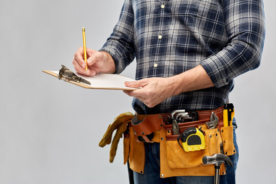 repair, construction and building - male worker or builder with clipboard, pencil and working tools on belt over grey background