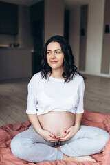 Photo of a pregnant brunette woman keeping her hands on belly while looking at camera. She is enjoying sunny morning in the bedroom.  Woman feeling her future baby through the belly.