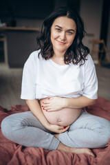 Photo of a pregnant brunette woman keeping her hands on belly while looking at camera. She is enjoying sunny morning in the bedroom.  Woman feeling her future baby through the belly.
