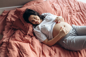 Pretty young brunette pregnant woman laying on a bed in a big light room. She is touching a belly by hands. Sun comes through the window. Bed linen of lush lava colour. Relaxing at Home. 