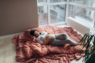 Pretty young brunette pregnant woman laying by the window in a big light room. She is touching a belly by hands. Sun comes through the window. Bed linen of lush lava colour. Relaxing at Home