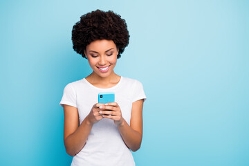 Photo of cheerful beautiful dark skin curly lady holding telephone hands chatting friends sending new pictures wear casual white t-shirt isolated blue color background