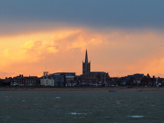 Fototapeta premium Orange sunset with clouds parting after a stormy day over St Nicholas Church on the seafront, Harwich, Suffolk, UK