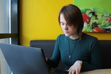 A girl with a calm expression sits, works at a laptop at a table in a cafe on a brown sofa against a yellow wall. He looks at the computer screen with his eyes closed