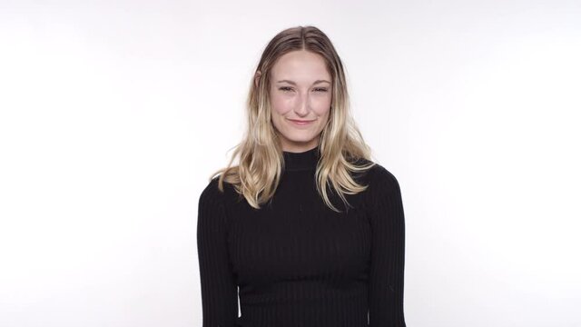 Young caucasian blonde woman smiles and shrugs on a white studio backdrop