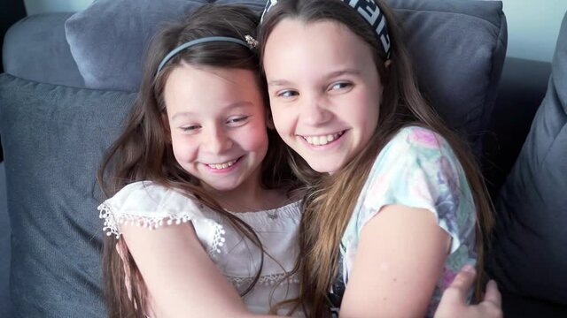 two teen girls hugging and laughing on the couch. Happy children lean together on the sofa. Slow motion