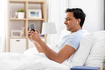 people, bedtime and rest concept - happy smiling indian man in earphones with smartphone lying in bed and listening to music at home