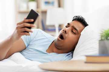 people, bedtime and rest concept - sleepy indian man with smartphone lying in bed at home and yawing