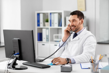 healthcare, medicine and people concept - smiling male doctor with clipboard calling on desk phone at hospital