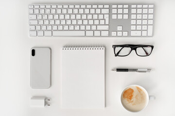 White keyboard, cup of coffee, mobile phone, blank notepad, pen, glasses and charger on the work desk. Flat lay. Knolling.