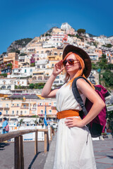 A girl in a white dress, sunglasses and a hat stands on the beach in Positano. View of houses and hotels in the background. Travel and vacation concept in Europe. With a backpack. Vertical photo