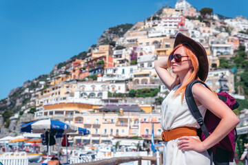 Obraz premium A girl in a white dress, sunglasses and a hat stands on the beach in Positano. View of houses and hotels in the background. Travel and vacation. Copy space. With a backpack on the back