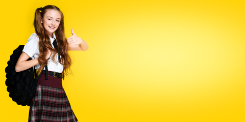 Elementary european schoolgirl with backpack showing thumb up and smiling on vibrant yellow background, pupil in uniform, banner format, education concept
