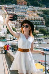 A woman in a white dress, sunglasses and a hat stands on the beach in Positano and makes selfie photo on smartphone. Travel and vacation concept in Europe. Vertical photo