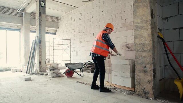 A male builder in an orange vest and an orange helmet approaches a pile of bricks and puts one brick in a common pile.