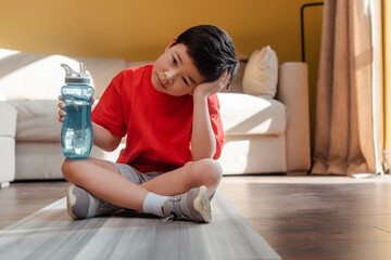 bored sportive asian boy with sports bottle sitting on fitness mat at home during quarantine