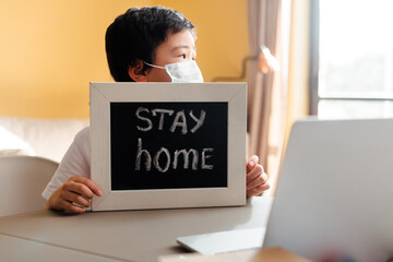 asian boy in medical mask holding board with stay home lettering in front of laptop during quarantine