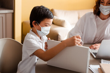 asian boy in medical mask studying with mother at home on quarantine