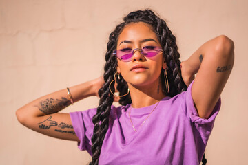 Urban session. Young dark-skinned woman with long braids wearing purple glasses on a plain cream-colored background, with serious look - obrazy, fototapety, plakaty