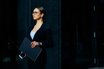 Confident female banker dressed in elegant stylish wear holding black folder in hands with mock up and looking away while standing near office.Copy space area for financial or business information