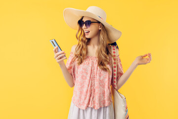 smiling young woman in a summer hat and sunglasses, holding a smartphone on an yellow background