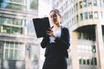 Prosperous young woman entrepreneur in formal apparel holding folder in hands standing on office building promotional background.Successful businesswoman in elegant wear with document's file