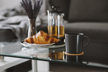 Herbal tea in a black mug and croissant served on a glass coffee table in living room