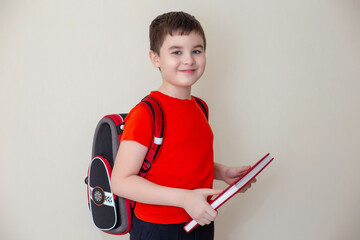 A smiling schoolboy in a red T-shirt, with a satchel behind his back, stands and holds a big book, looks at the camera