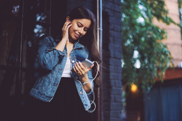 Good looking young woman with brunette hair watching video in social networks in earphones connected to modern smartphone device while strolling in urban setting enjoying leisure time