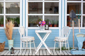 White table and chair on veranda of house. Yard with gardening tools. Facade blue Cozy home with a beautiful garden in summer. Beautiful farmhouse with wicker baskets and green plants on terrace.	
