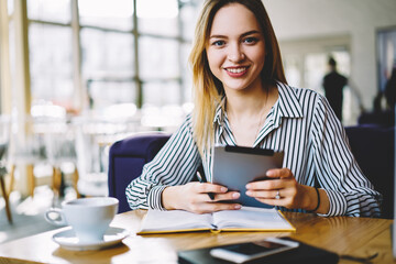 Portrait of cheerful blonde business woman checking financial documentation and accountings using application on touchpad, beautiful female entrepreneur making plannings of working task in notepad