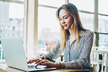 Fototapeta premium Cheerful female copywriter keyboarding text on digital netbook and searching information on internet websites for productive work.Positive freelancer updating software on laptop device sitting indoors