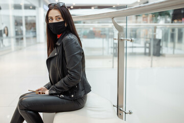 Obraz premium Girl with medical black mask and mobile phone in a shopping center. Coronavirus pandemic. A woman with a mask is standing in a shopping center. A girl in a protective mask is shopping at the mall